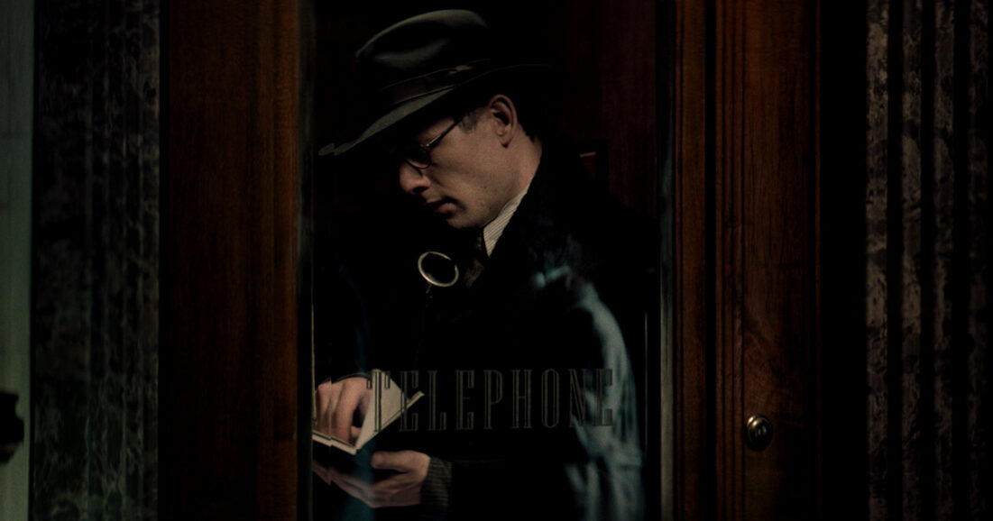 Na zdjęciu mężczyzna w kapeluszu stojący w drewnianej budce telefonicznej i przeglądający zeszyt.