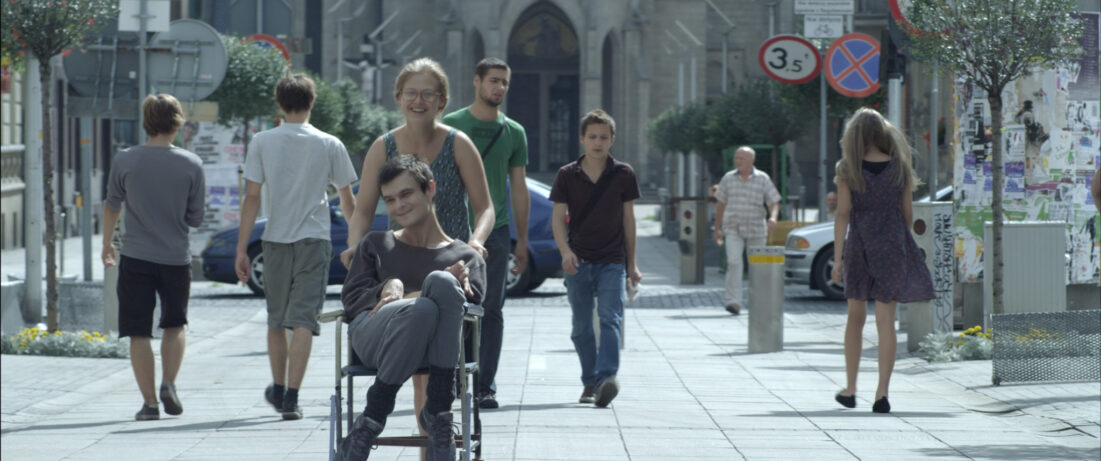 Na zdjęciu młoda kobieta w okularach prowadząca wózek inwalidzki, na którym siedzi młody mężczyzna.