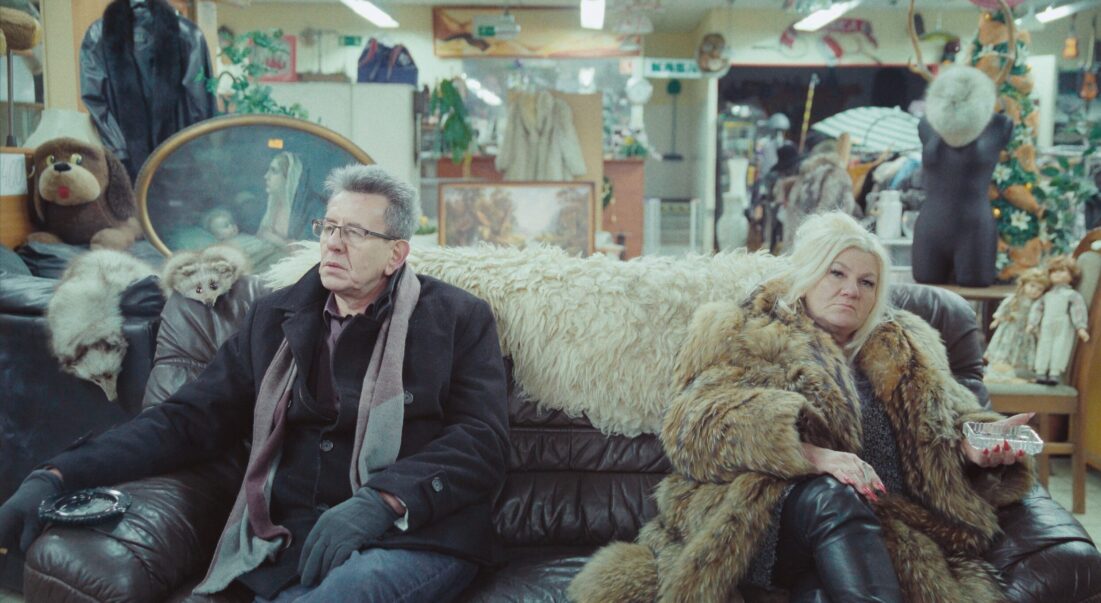 Na zdjęciu mężczyzna w płaszczu i kobieta w futrze siedzący na kanapie w pomieszczeniu wypełnionym przedmiotami.