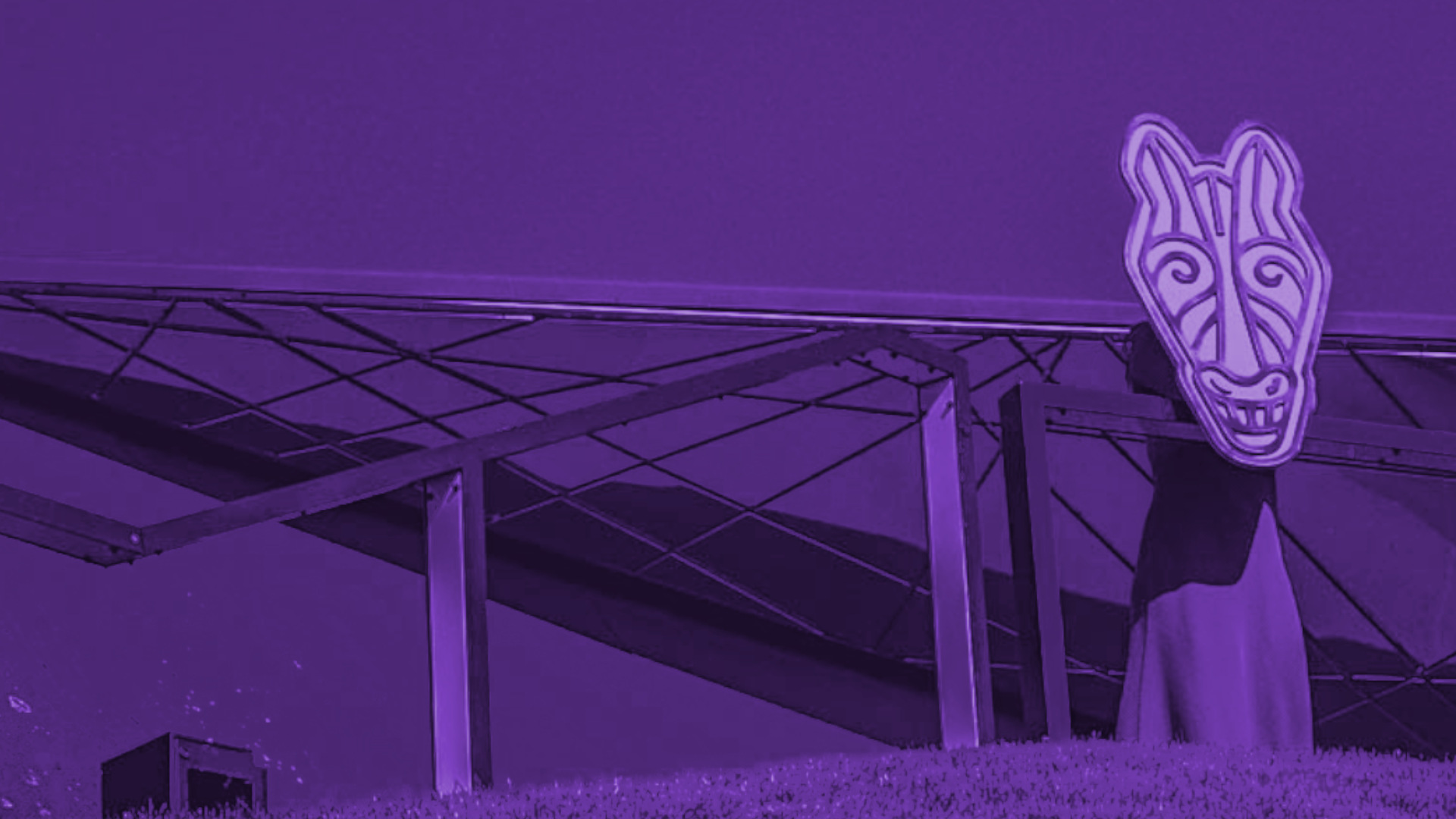 Człowiek stojący na tle metalowej konstrukcji trzymający papierową graficzną głowę konia w miejscu swojej głowy.